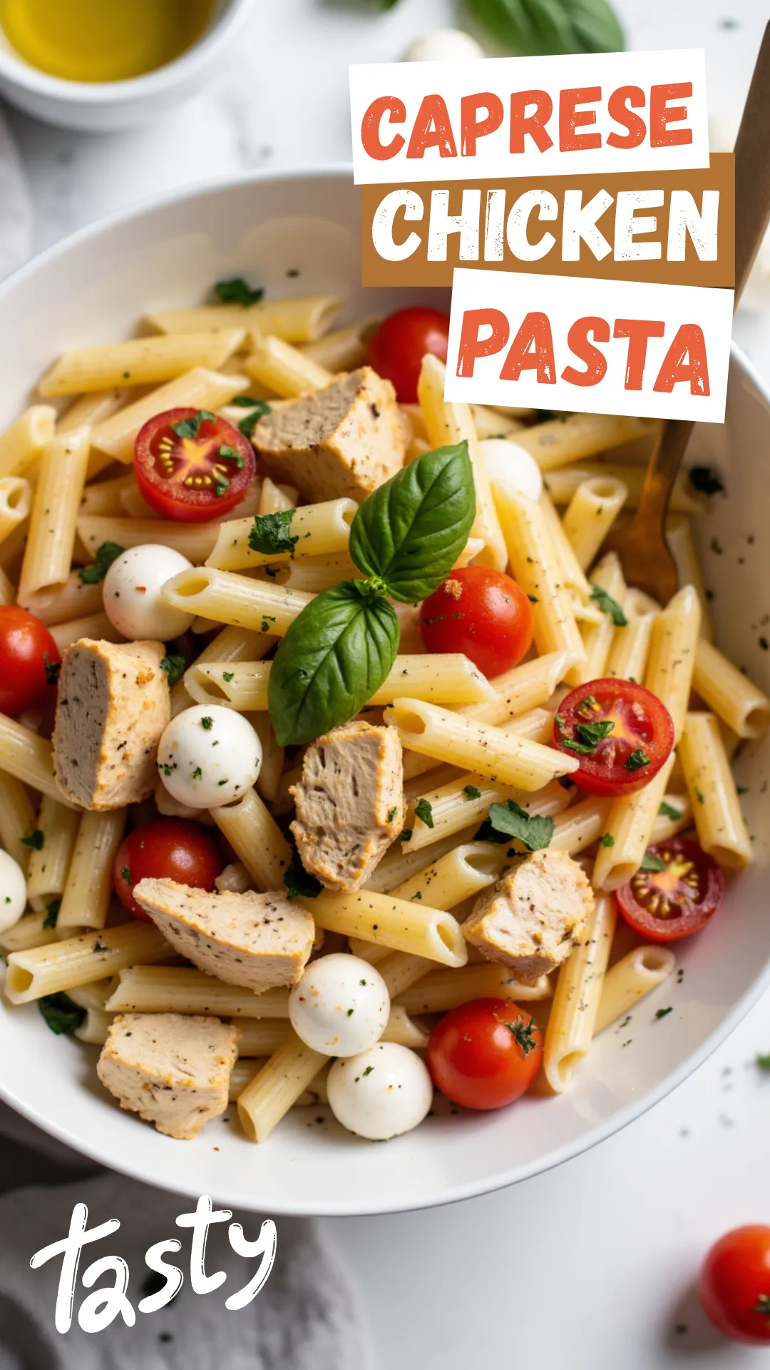 Overhead shot of a white bowl filled with penne pasta, grilled chicken chunks, cherry tomatoes, mozzarella pearls, and fresh basil, drizzled with olive oil, bright natural light